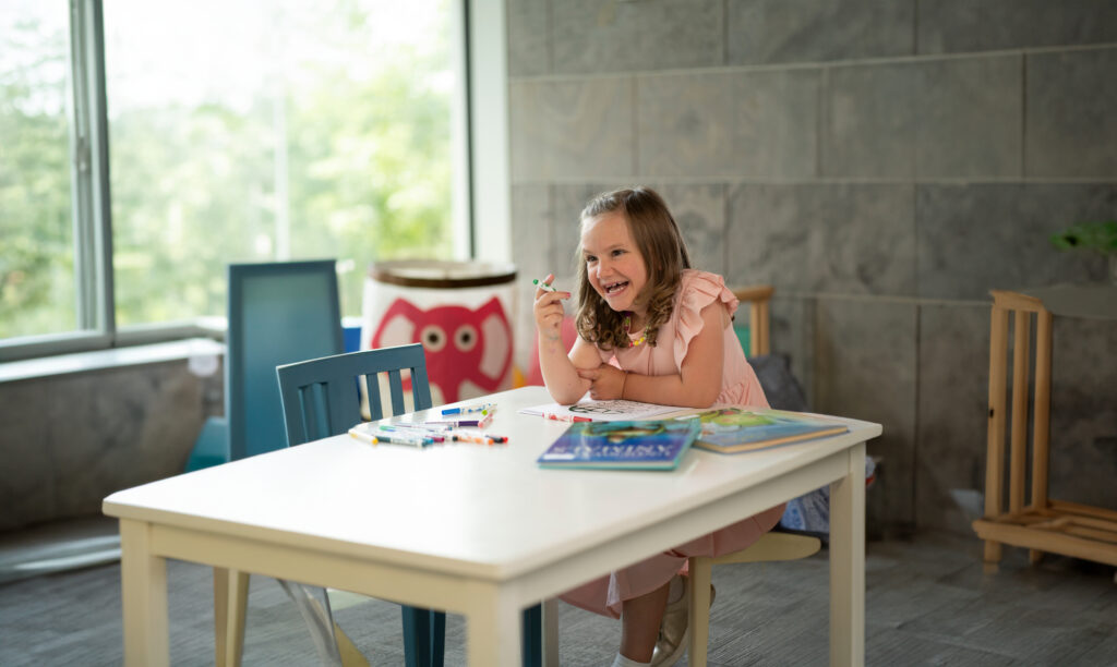 A girl, six years old, sits at the table and colours. She smiles at someone off camera.
